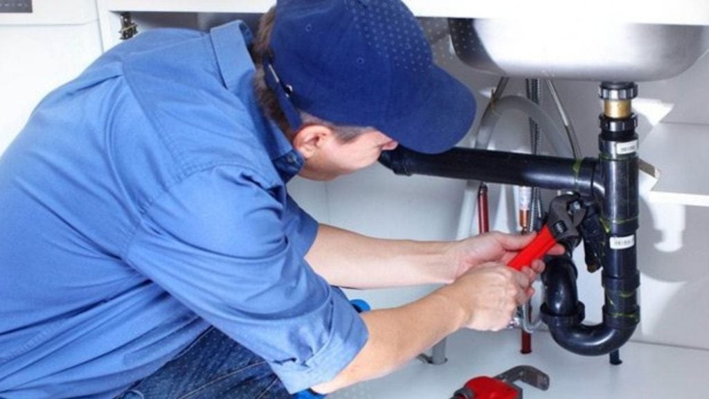 A plumber in a blue shirt and cap uses a pipe spanner to fix a blocked sink under the counter. - The Handyman Services York Region & Simcoe Region