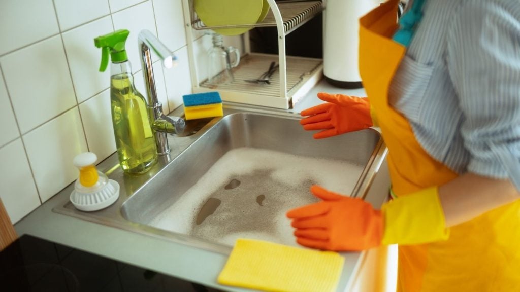How to fix a clogged sink. Person wearing orange gloves and a yellow apron stands by a kitchen sink filled with soapy water, surrounded by cleaning supplies—perfect for learning how to fix a blocked sink while tackling household chores. - The Handyman Services York Region & Simcoe Region