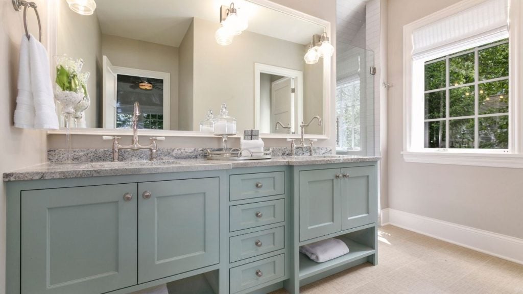 A bathroom with a double-sink vanity in light blue, granite worktop, large mirror, glass shower, towel, and a window letting in natural light. - The Handyman Services York Region & Simcoe Region