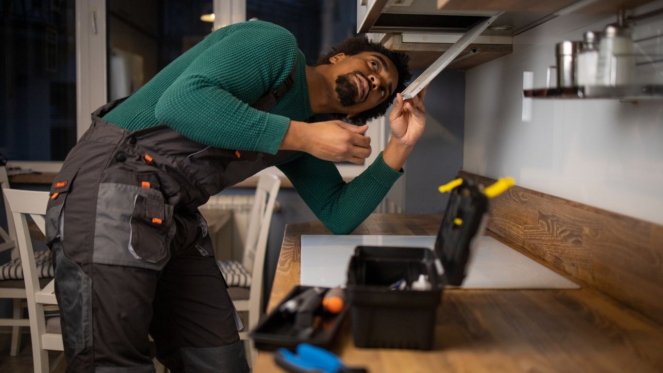 A man in work overalls repairs a kitchen cupboard using tools from a black toolbox on the worktop. - The Handyman Services East Gwillimbury